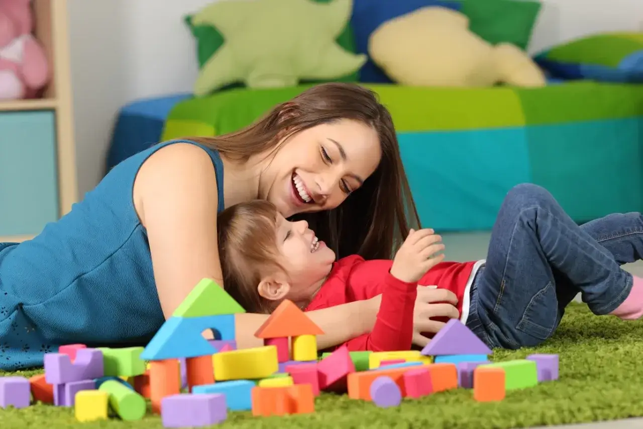 Uśmiechnięta opiekunka bawi się z dzieckiem klockami. Zastanawiasz się, ile zarabia opiekunka dla dziecka?