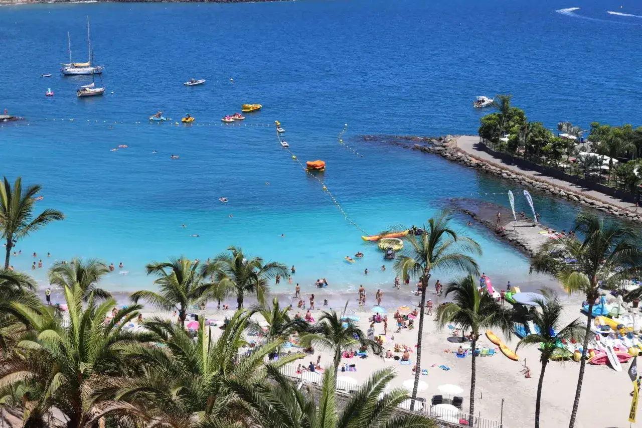 Słoneczna plaża na Gran Canarii, z turkusową wodą, palmami i ludźmi odpoczywającymi na piasku. Idealne miejsce na wakacje, jak z przewodnika po Gran Canarii.