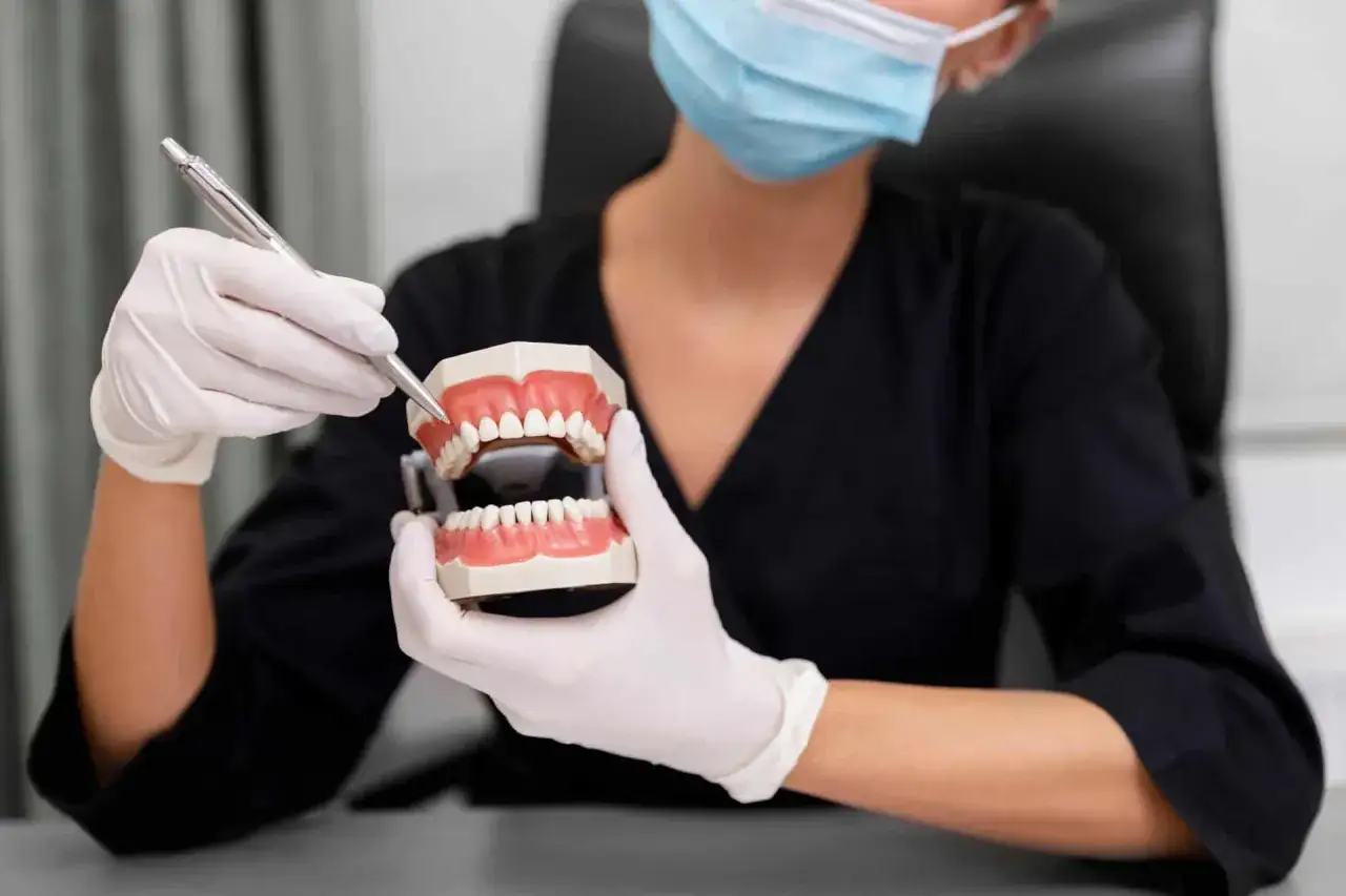 Dentysta w masce i rękawiczkach pokazuje model szczęki, wyjaśniając, kto robi protezy zębowe.