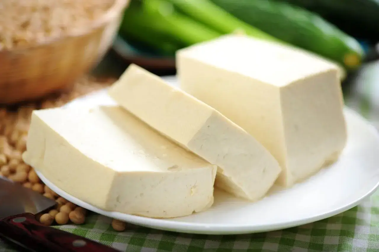 Bloki tofu, które powstają z ziaren soi, leżą na talerzu. W tle widać miskę z ziarnami soi i zielone warzywa.