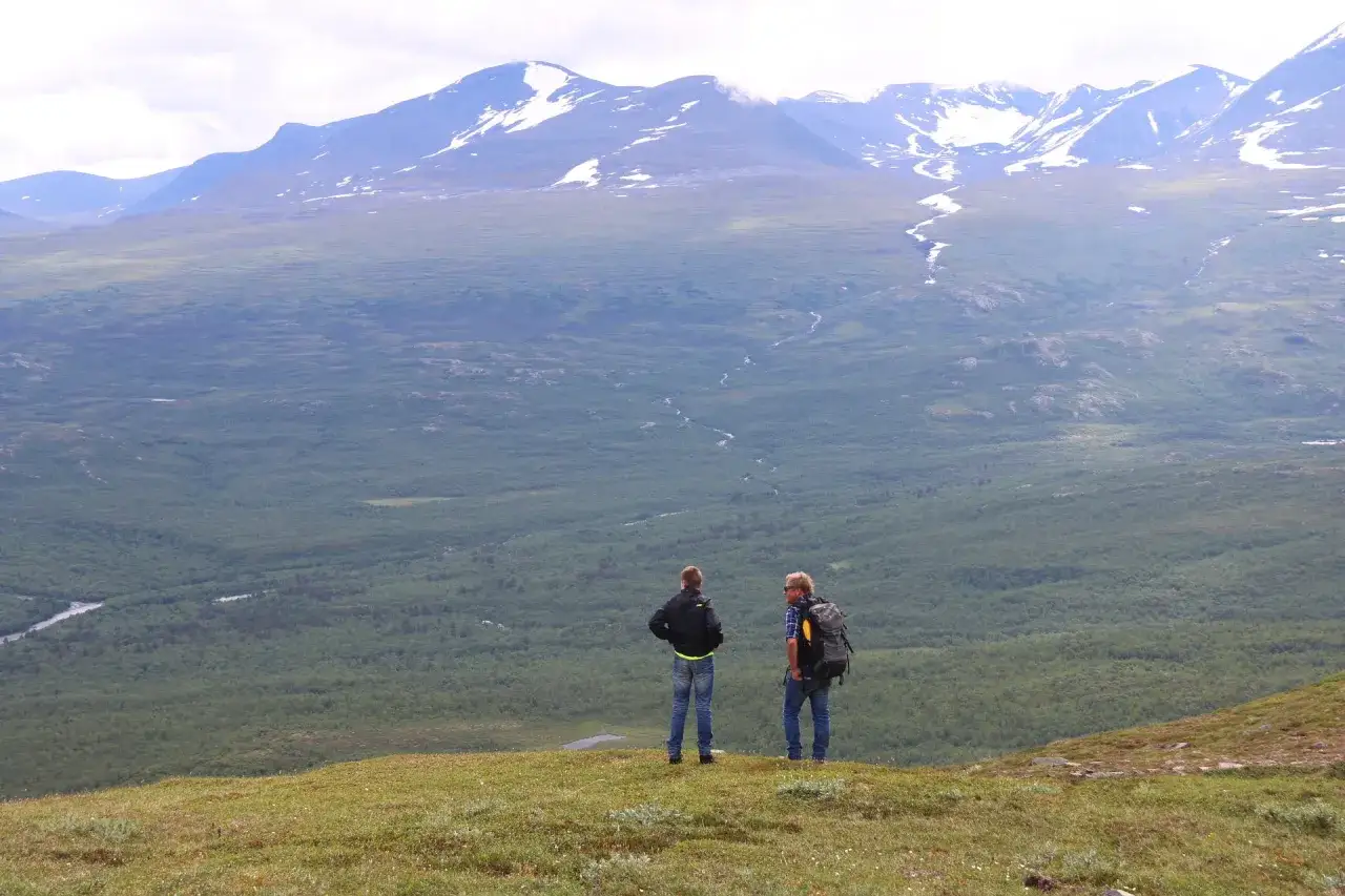 Tv&aring; vandrare njuter av den vidstr&auml;ckta vyn &ouml;ver fj&auml;llandskap i Abisko sommar. Sn&ouml;kl&auml;dda bergtoppar reser sig mot en molnig himmel.