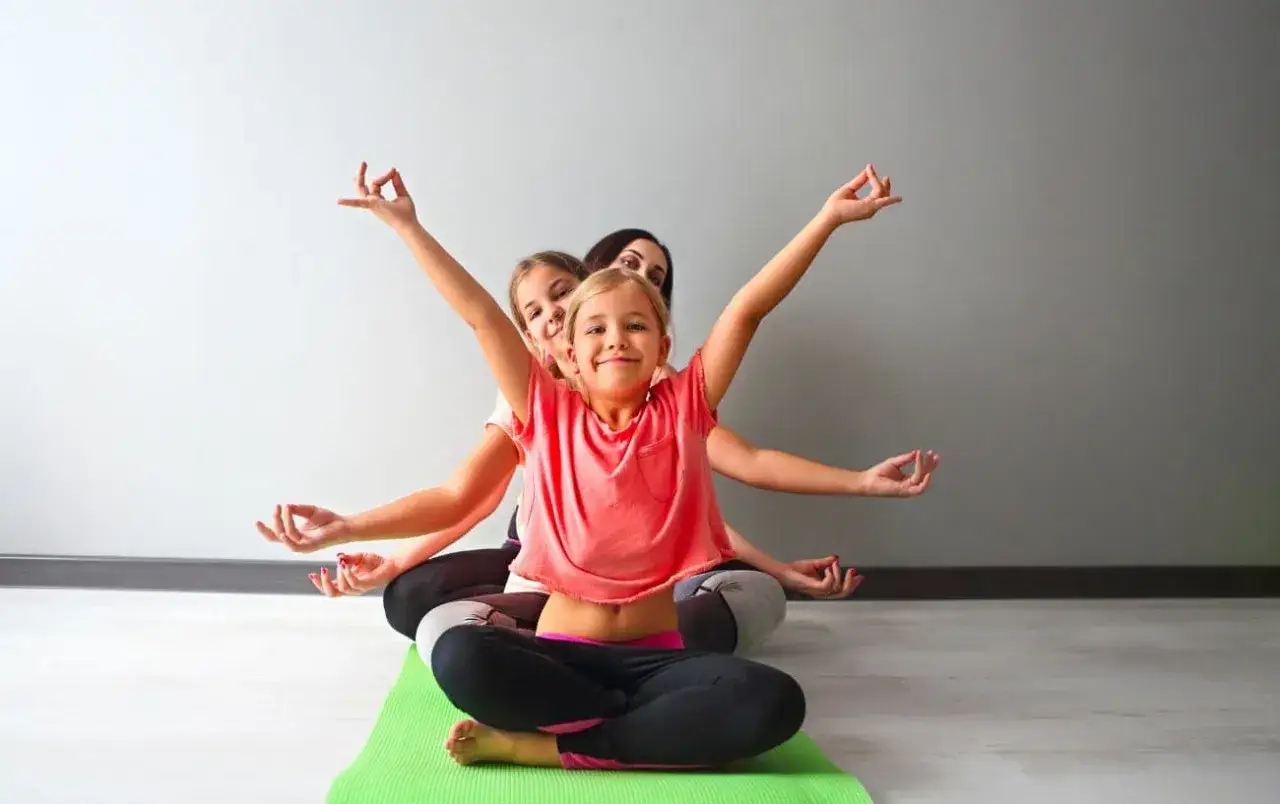 Drei M&auml;dchen &uuml;ben Yoga. Sie sitzen im Schneidersitz auf einer gr&uuml;nen Matte und machen eine Handgeste. Yoga-geschichten f&uuml;r kinder zum mitmachen.