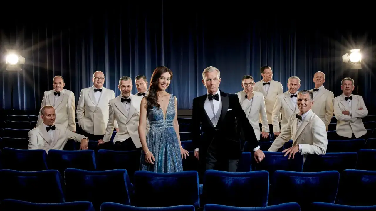 Max Raabe och hans Palast Orchester i Stockholm, kl&auml;dda i eleganta vita kostymer och en glittrig bl&aring; kl&auml;nning, poserar framf&ouml;r en m&ouml;rkbl&aring; rid&aring;.