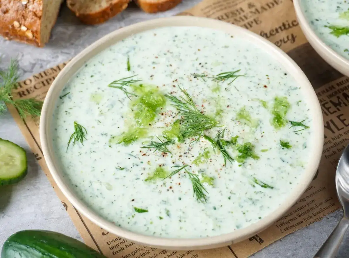 Eine Schüssel cremige kalte Gurkensuppe mit Dill und zerdrückten Gurken, garniert mit Pfeffer. Dazu gibt es Brot.