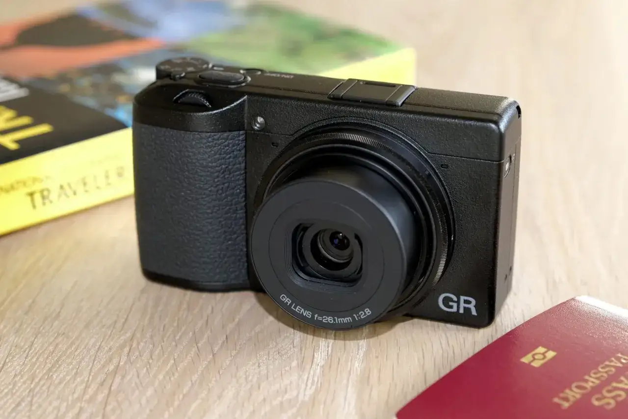 En svart Ricoh GR III X-kamera ligger p&aring; ett tr&auml;bord bredvid en resebok och ett pass. Perfekt f&ouml;r att f&aring;nga dina &auml;ventyr.