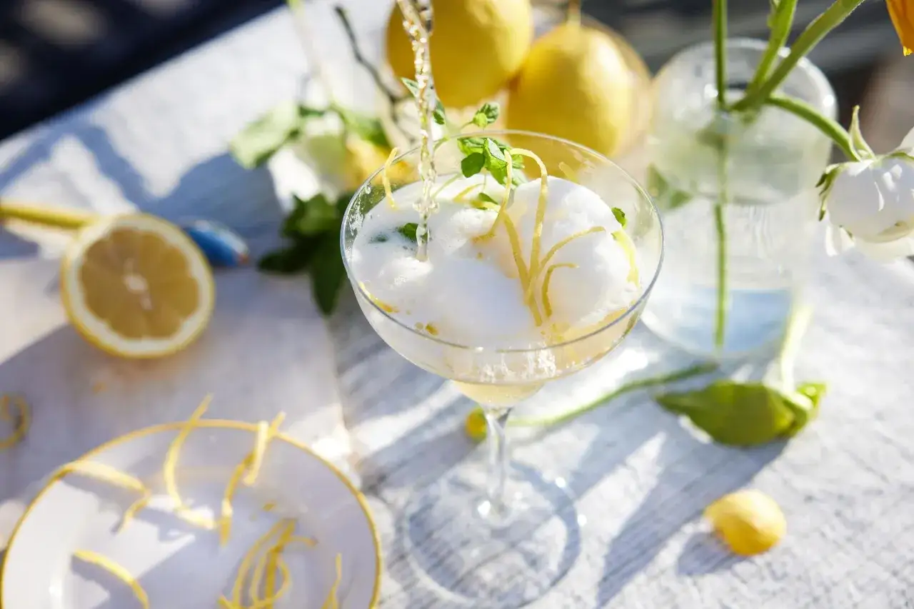 En bubblande alkoholfri drink med citronskal och mynta hälls upp i ett glas. Citroner och blommor i bakgrunden skapar en somrig känsla.