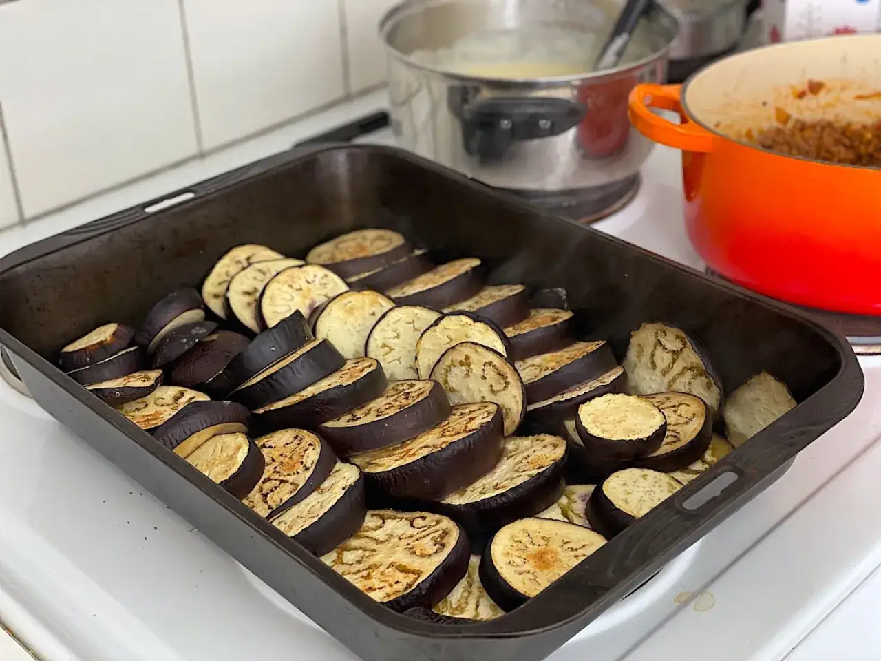 Skivad aubergine redo för ett moussaka recept. I bakgrunden syns grytor med mat.