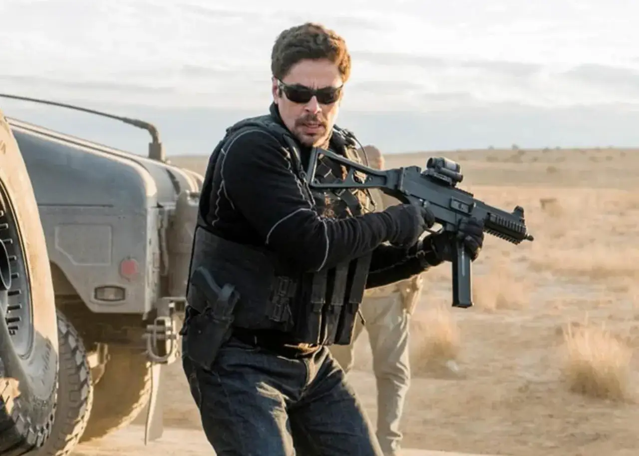 Sicario 3: Alejandro w czarnym stroju, z pistoletem i karabinem, gotowy do akcji na pustyni.