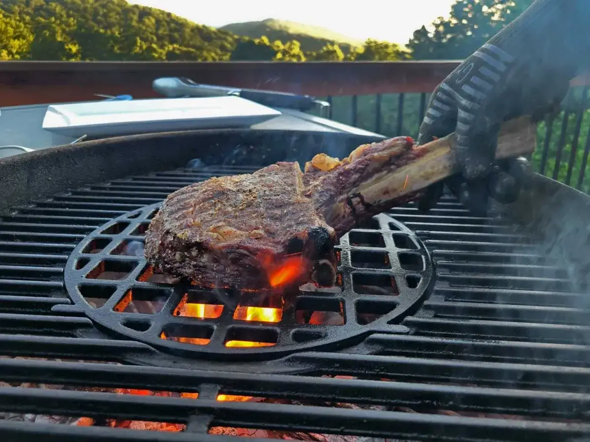 En hand med grillhandske vänder på en saftig tomahawk-biff över glödande kol.