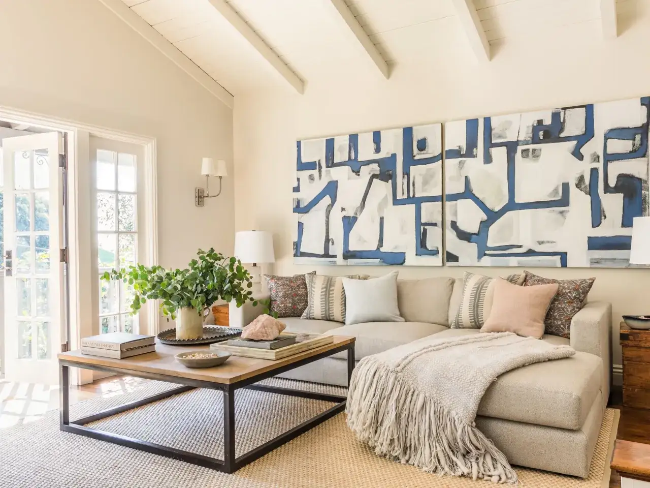 Sof&aacute; beige con cojines y manta, mesa de centro de madera y metal, arte abstracto azul y blanco. Elegantes combinaciones con beige.