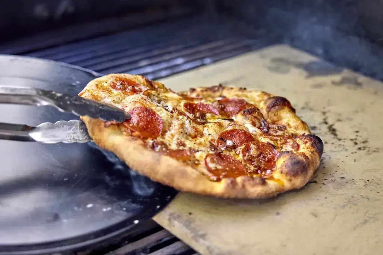 Eine k&ouml;stliche Pizza mit Salami wird mit einer Zange vom Pizzastein auf dem Grill gehoben.