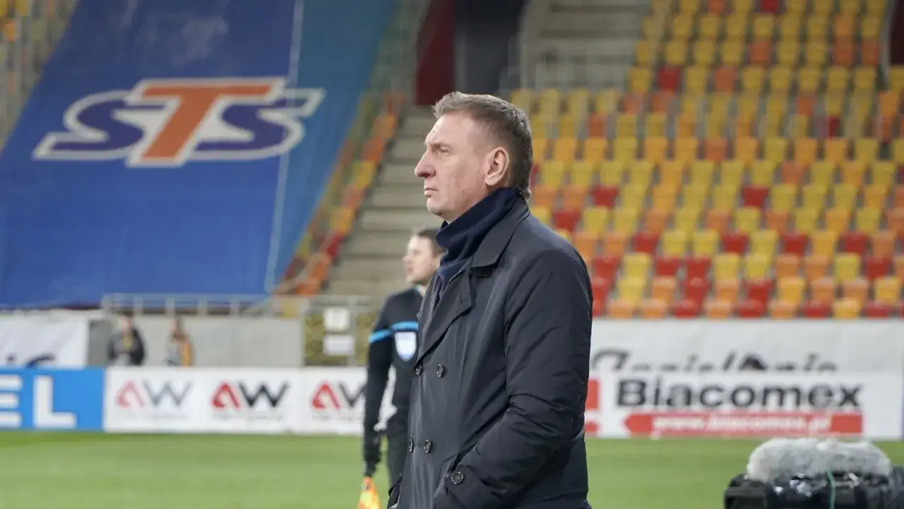 Kamil Kieres, trener w ciemnym płaszczu, stoi na tle stadionu z widownią w żółto-czerwonych barwach.