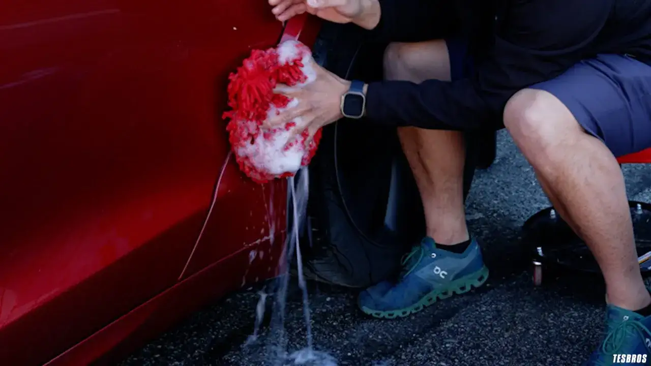 A person demonstrates how to power wash a car, using a red, foamy mitt to scrub the side of a shiny red vehicle.