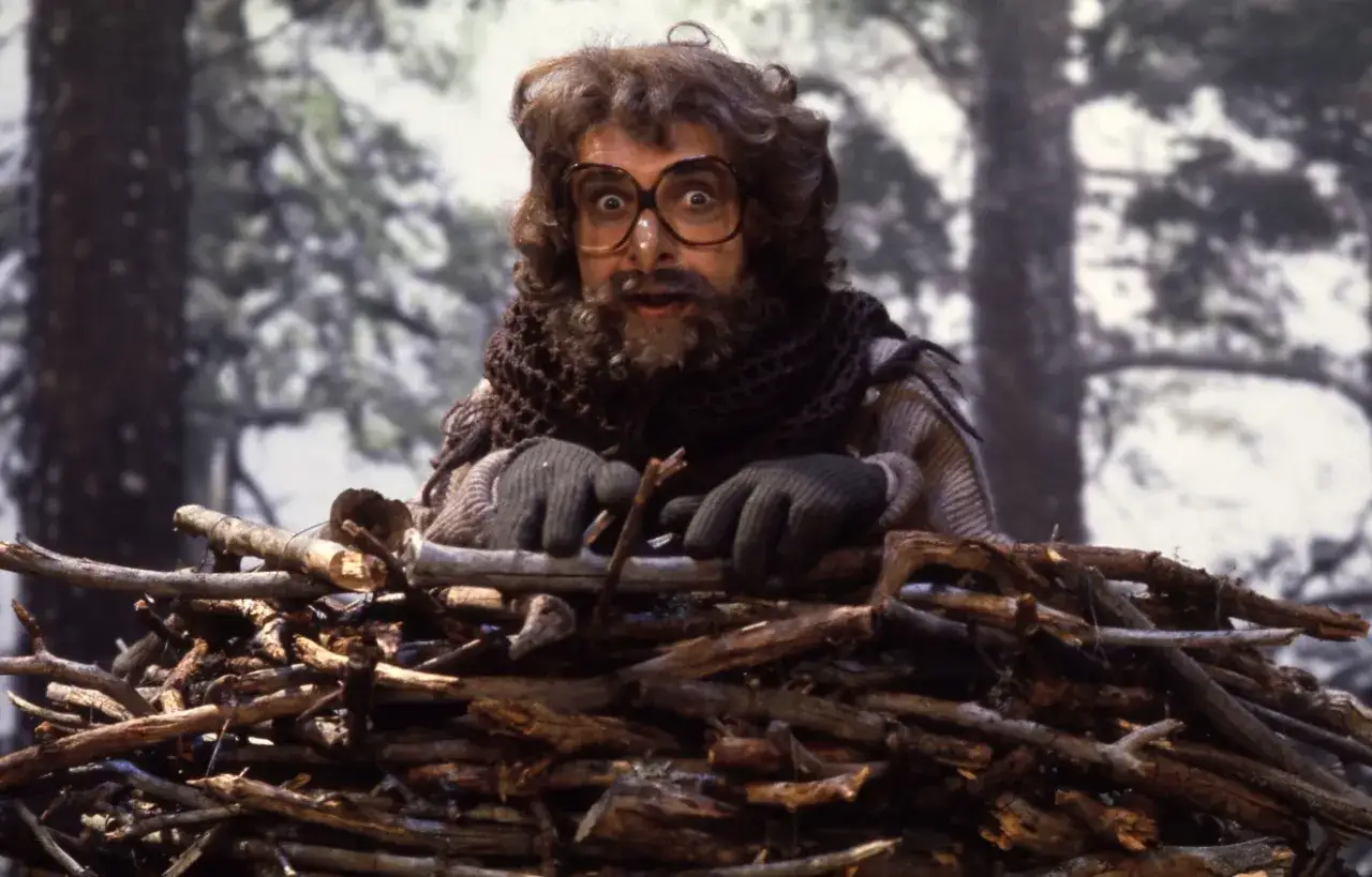 Ugglan Helge, med stora glas&ouml;gon och yvig frisyr, tittar f&ouml;rv&aring;nat fram bakom en h&ouml;g med grenar i skogen.