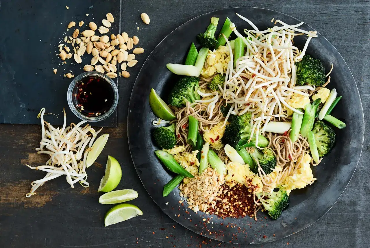 En tallrik med pad see ew, nudlar, broccoli, böngroddar, ägg och lime. Jordnötter och chilisås vid sidan.