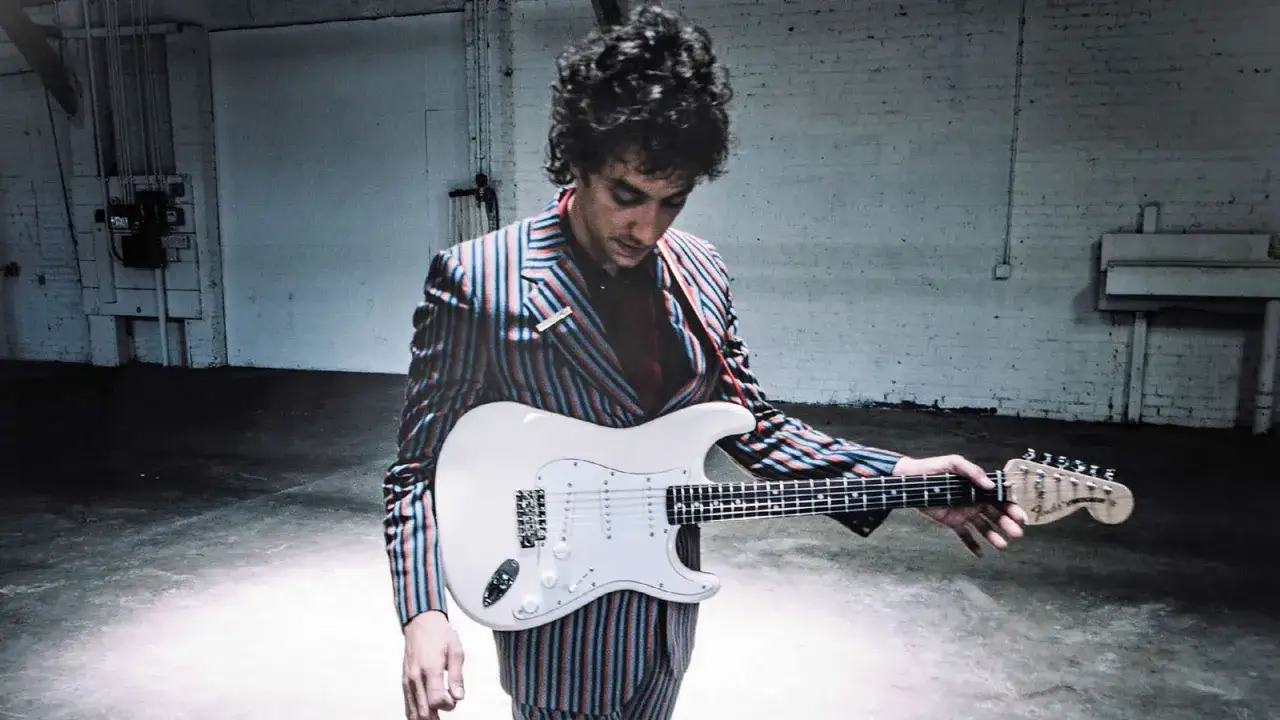 Albert Hammond Jr. w stylowym garniturze z prążkami trzyma białą gitarę elektryczną w opuszczonym magazynie.
