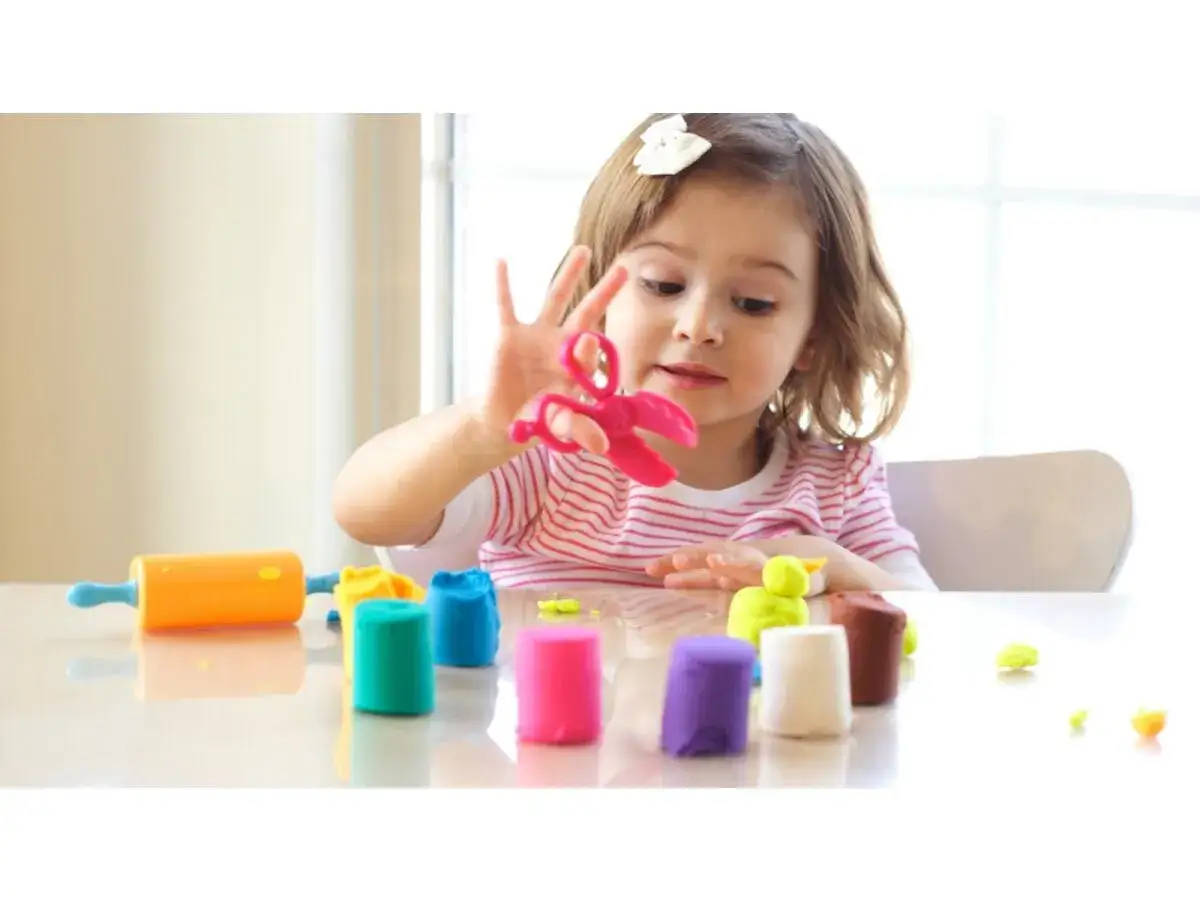 Bambina gioca con pasta modellabile colorata, forbici rosa e mattarello arancione. Giochi di manipolazione Montessori per sviluppare creatività e manualità.