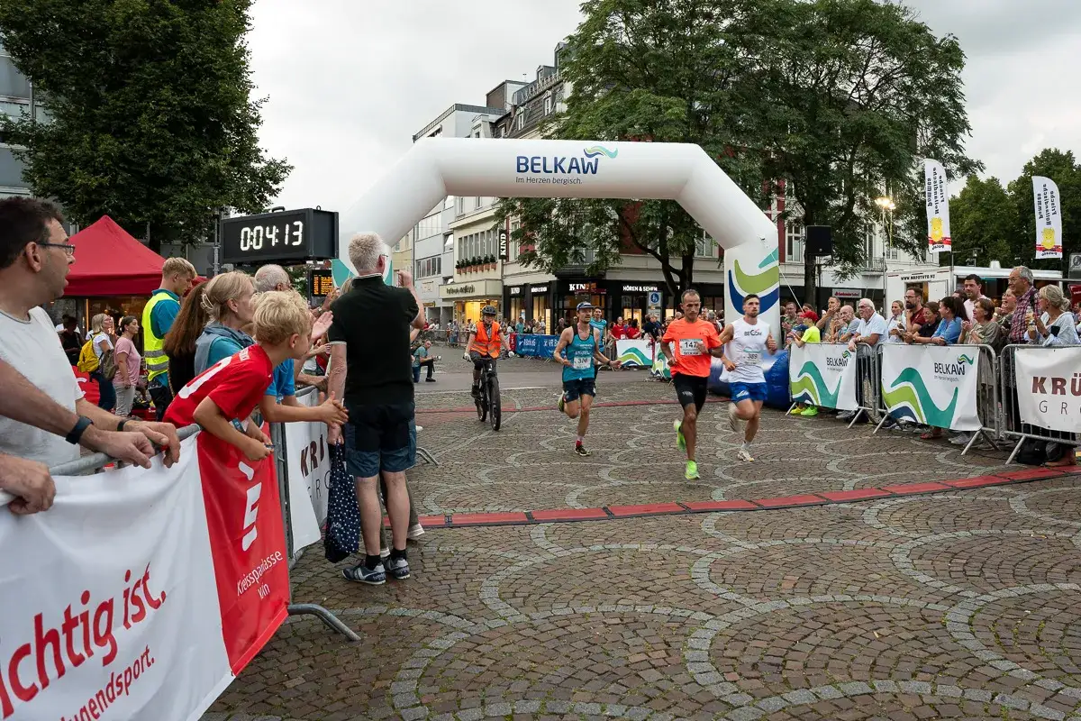 L&auml;ufer &uuml;berqueren die Ziellinie beim Stadtlauf Bergisch Gladbach unter dem BELKAW-Bogen. Zuschauer feuern sie an.