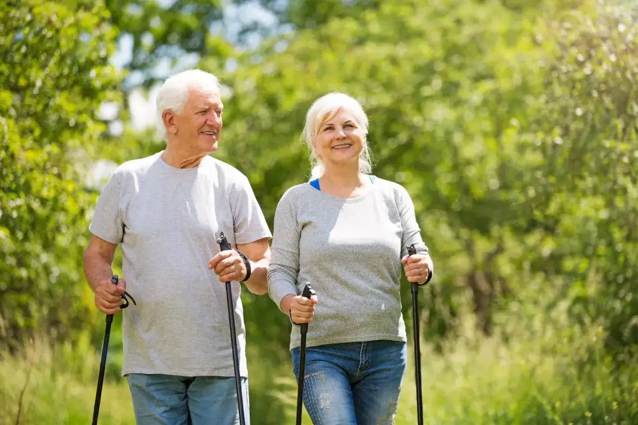 Uśmiechnięta para seniorów cieszy się aktywnością na świeżym powietrzu, uprawiając **nordic walking** wśród zieleni.