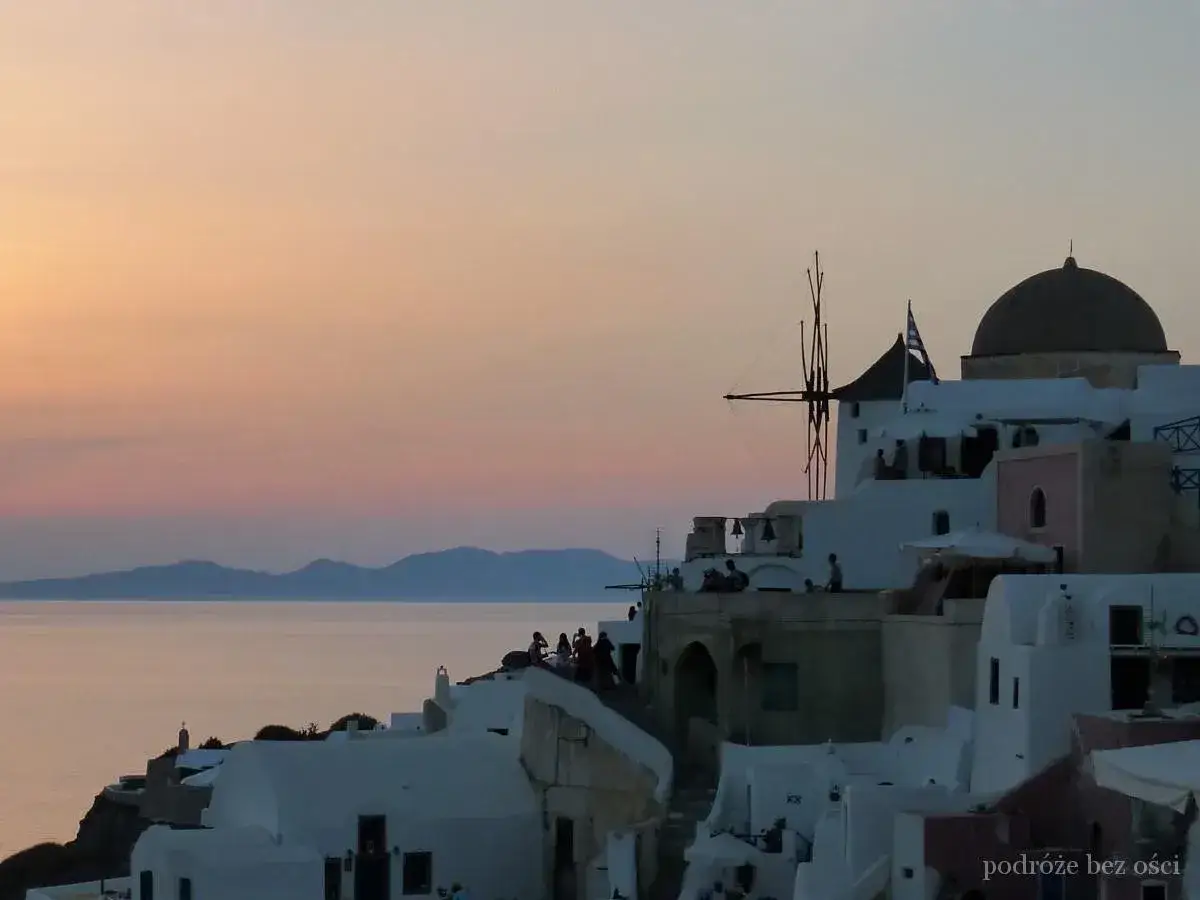 Zach&oacute;d słońca na Santorini w maju. Białe domy, wiatrak i kopuła kościoła na tle pastelowego nieba i morza. Idealna pogoda na zwiedzanie.