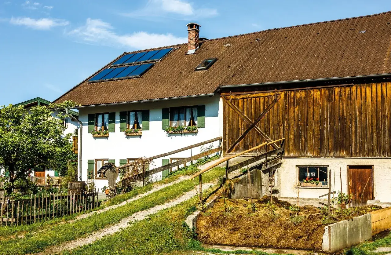 Ein Bauernhof mit Solaranlagen auf dem Dach und einem großen Holztor. Der **Altenteiler Landwirtschaft** genießt die Sonne.
