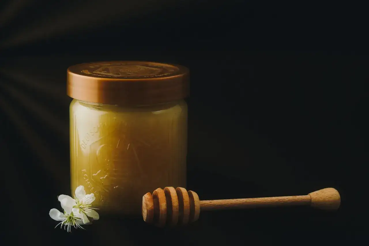 Słoik z płynnym, nieskrystalizowanym miodem naturalnym, drewniana chochla i biały kwiatek na ciemnym tle.