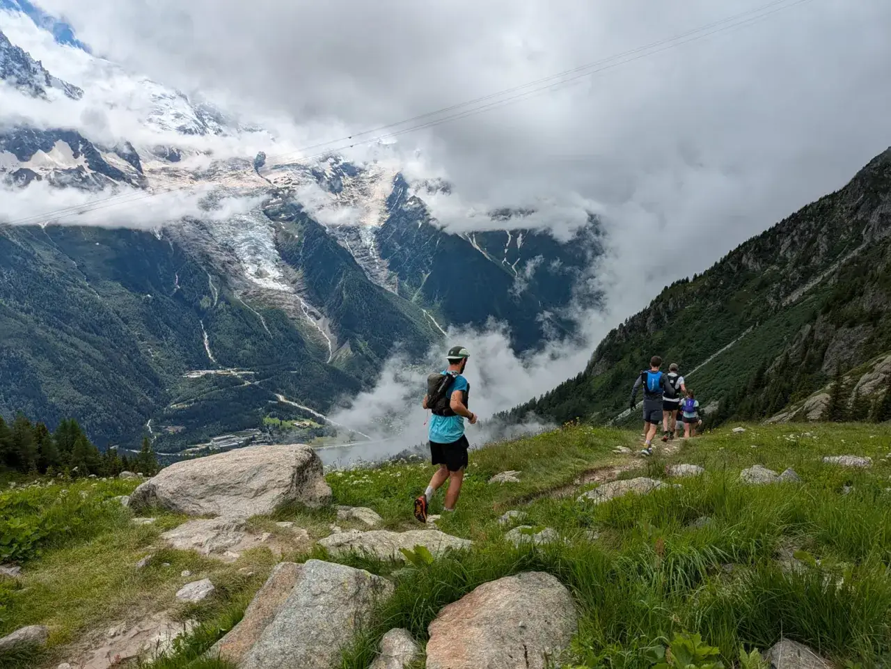 Coureurs en entra&icirc;nement pour l'UTMB, traversant un sentier de montagne verdoyant avec des glaciers et des nuages en arri&egrave;re-plan.