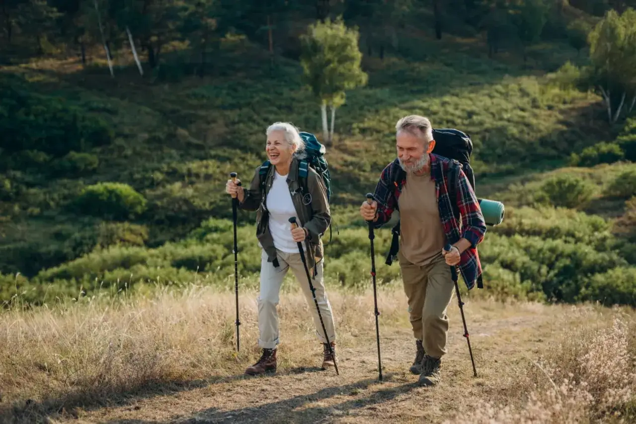 Uśmiechnięta para seniorów wędruje po wzgórzu, pokazując, jak chodzić z kijkami.