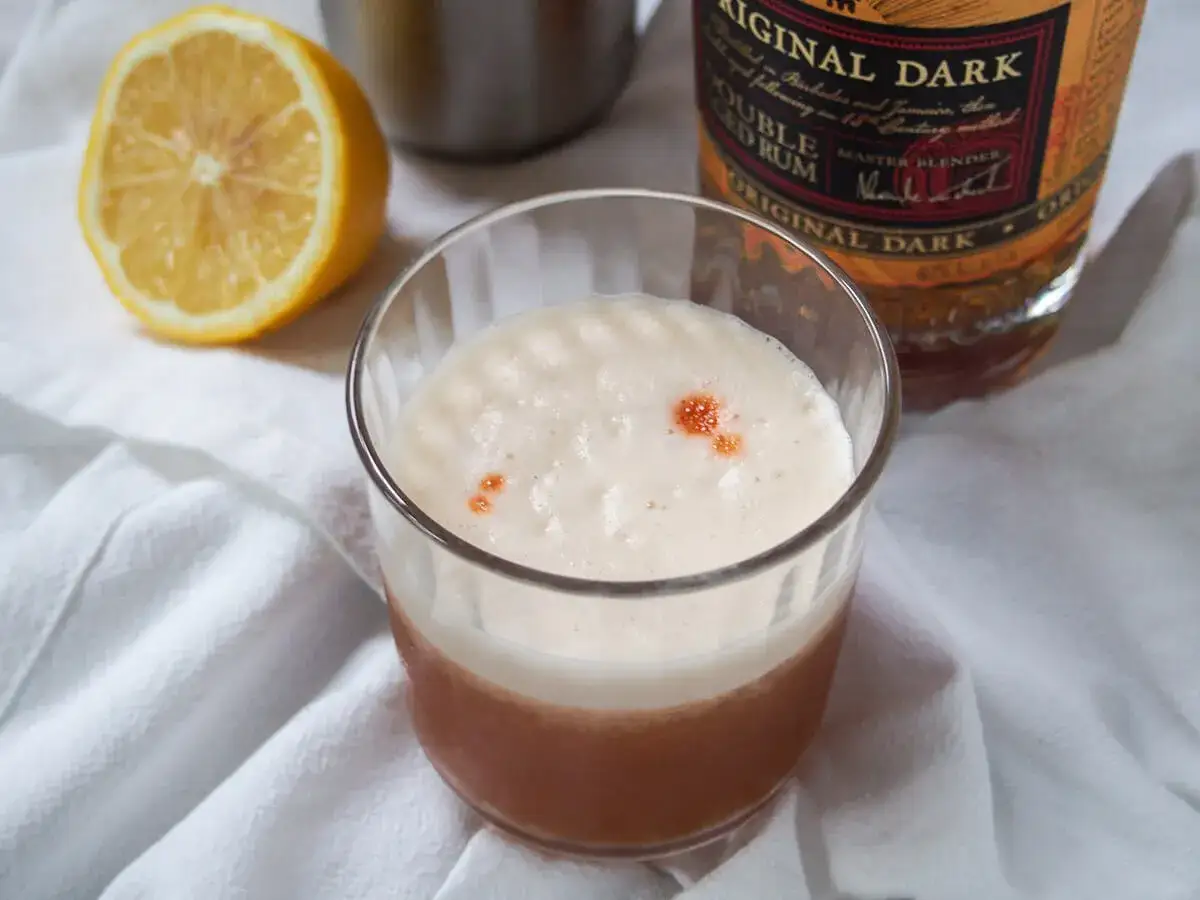 Ein cremiger Rum Sour mit Zitronenspalte und einer Flasche dunklem Rum im Hintergrund.