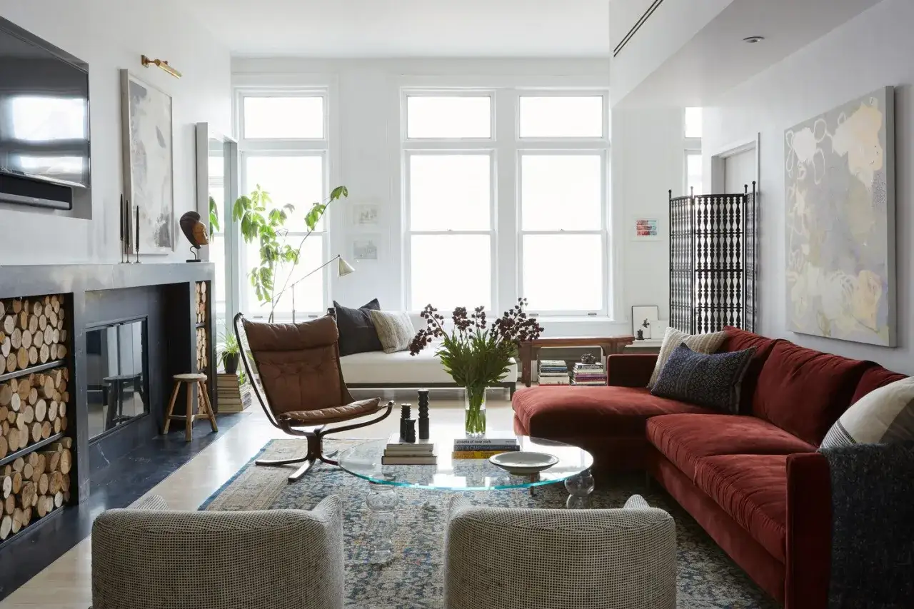 Ein Wohnzimmer mit einem tiefroten Samtsofa, einem Ledersessel, einem Couchtisch aus Glas und einem Kamin mit Holzstapeln.