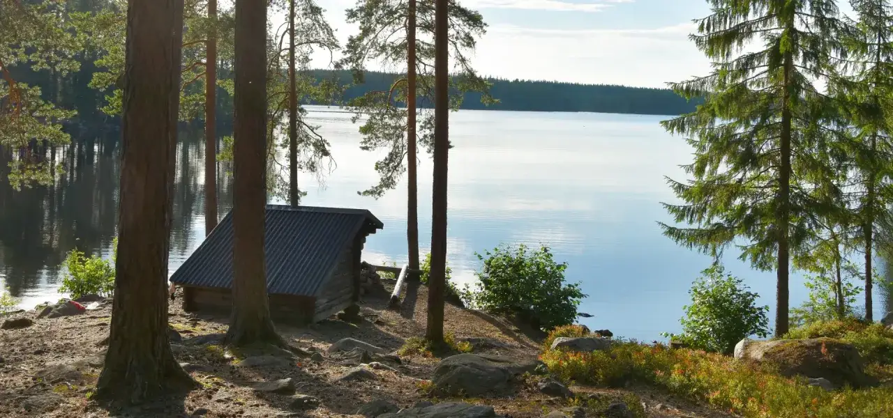 En liten stuga vid en sjö, omgiven av tallar. Kanske en del av Malingsbo herrgård, en idyllisk plats.