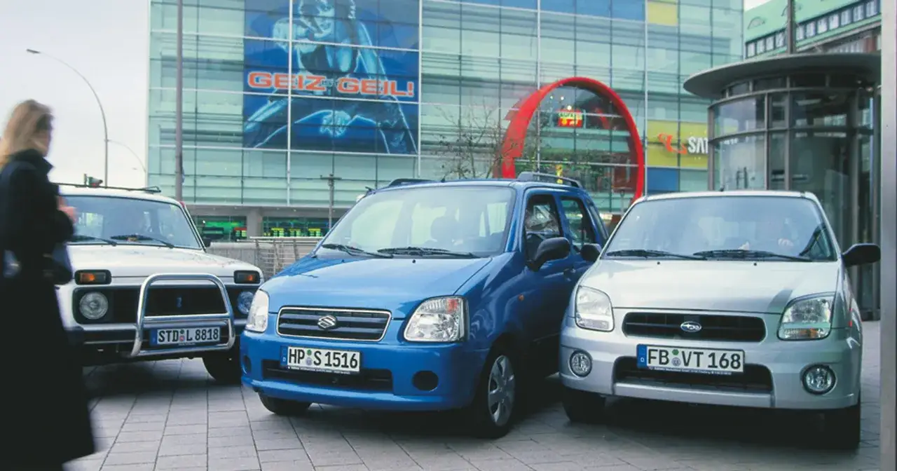 Niebieski Subaru Justy obok srebrnego, z budynkiem w tle.