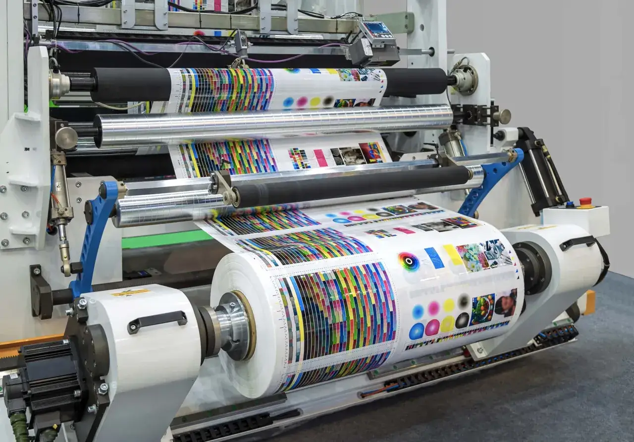 M&aacute;quina de impresi&oacute;n industrial en acci&oacute;n, mostrando el proceso de **impresi&oacute;n litogr&aacute;fica** con rollos de papel llenos de coloridos patrones y pruebas de impresi&oacute;n.