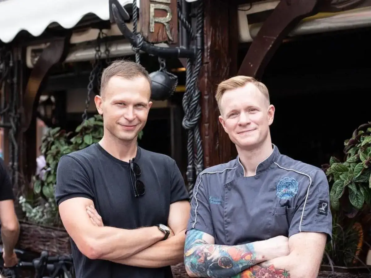 Dwóch mężczyzn, jeden w czarnej koszulce, drugi w szarym fartuchu, pozuje przed restauracją. To część obsady filmu