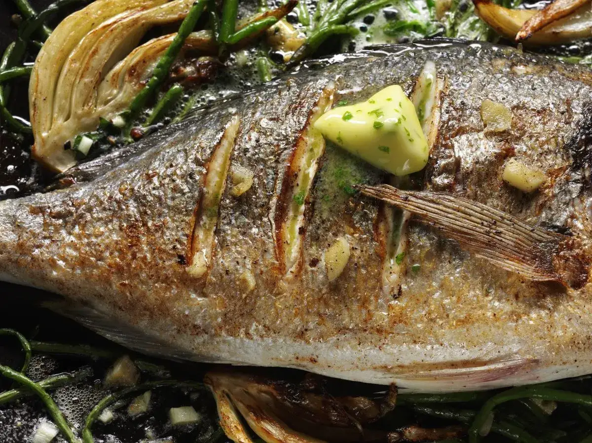 Złocisty sea bass pieczony z czosnkiem, koprem i masłem, podany z fenkułem i zielonymi warzywami.
