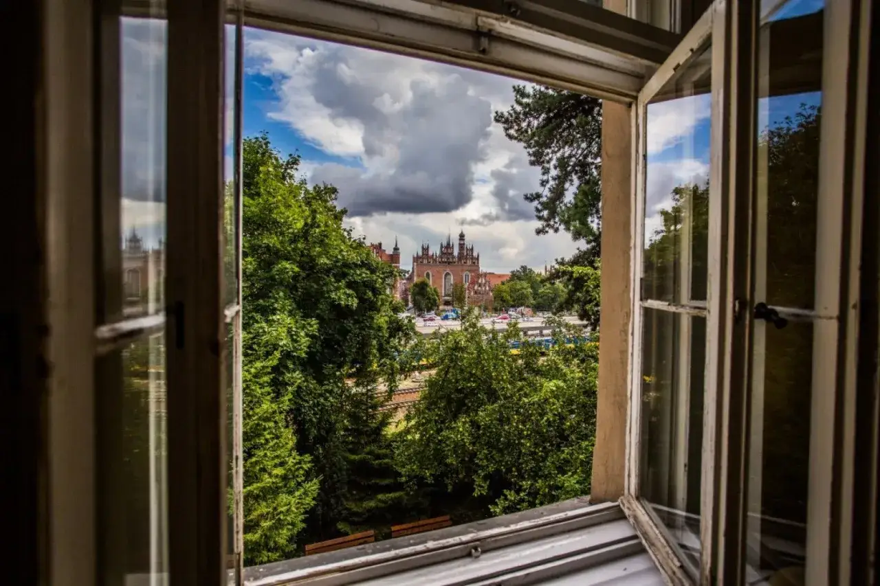 Widok z okna na zabytkową Biskupią Górkę, zdominowaną przez ceglaną architekturę i bujną zieleń.