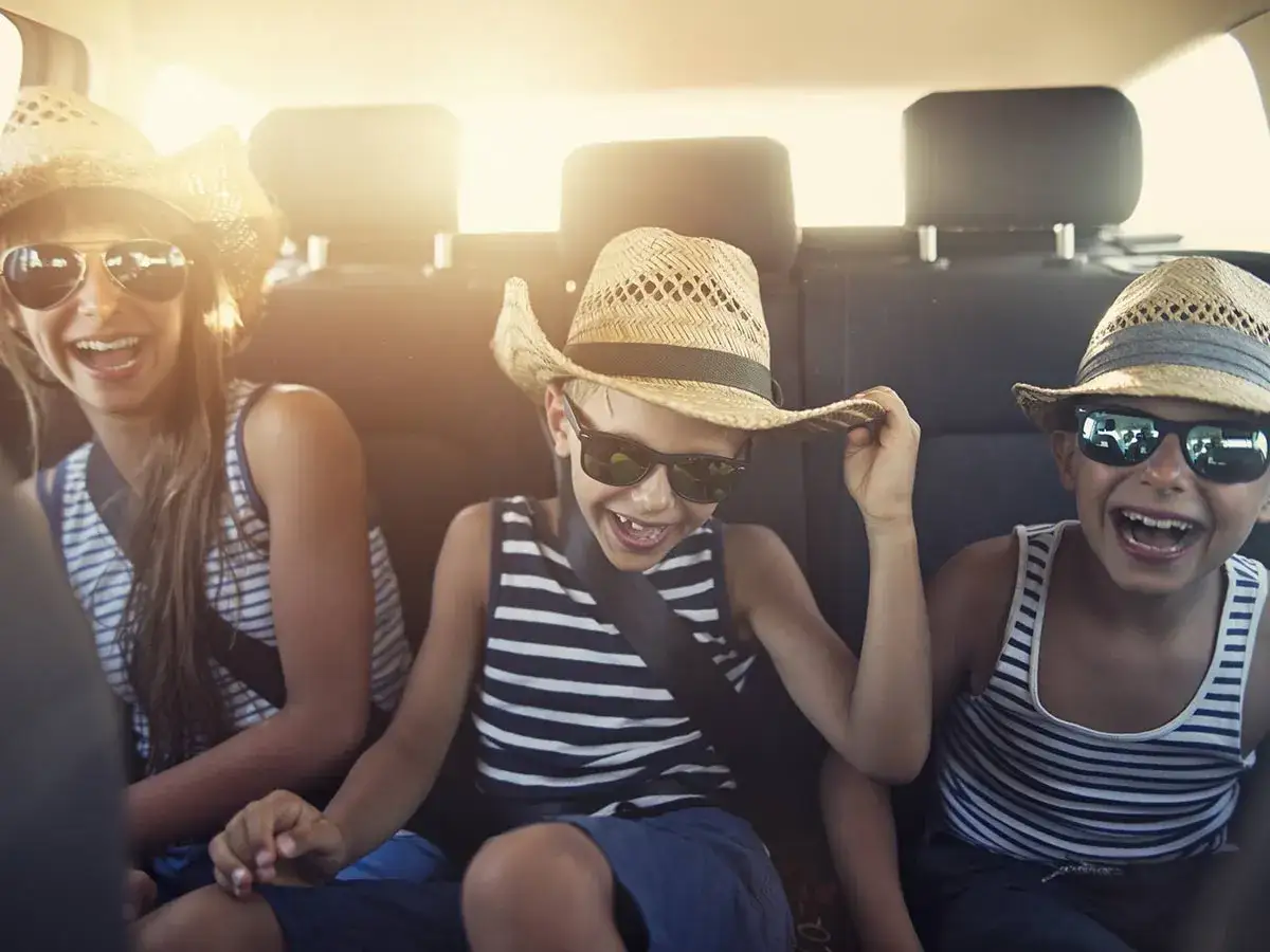 Drei lachende Kinder mit Strohhüten und Sonnenbrillen im Auto. Perfekt für Reisespiele mit Kindern!