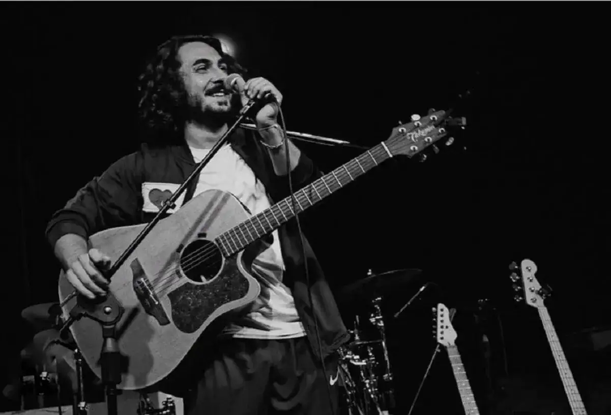 Un postino cantante con chitarra e microfono sul palco, circondato da strumenti musicali.