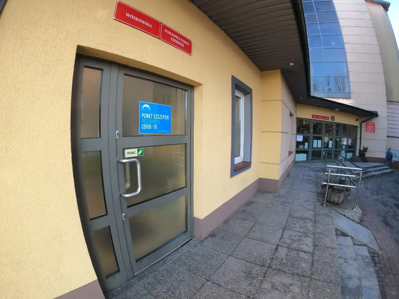 Drzwi do punktu szczepień przeciw COVID-19. Na drzwiach widnieje niebieska naklejka z napisem 