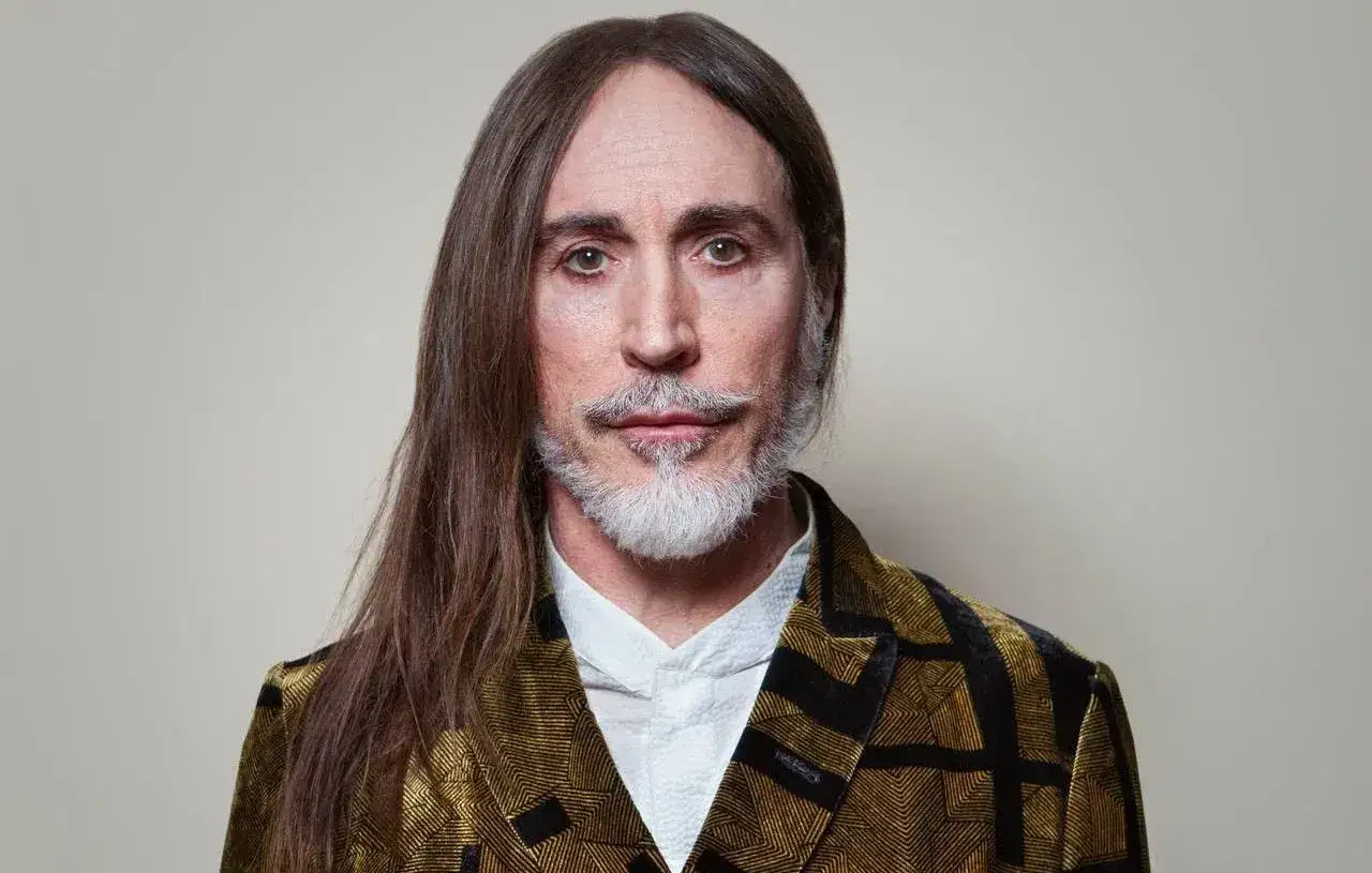 Ritratto di un uomo con lunghi capelli castani, barba e baffi grigi, indossa una giacca dorata e nera. Uno dei cantanti di Milano.