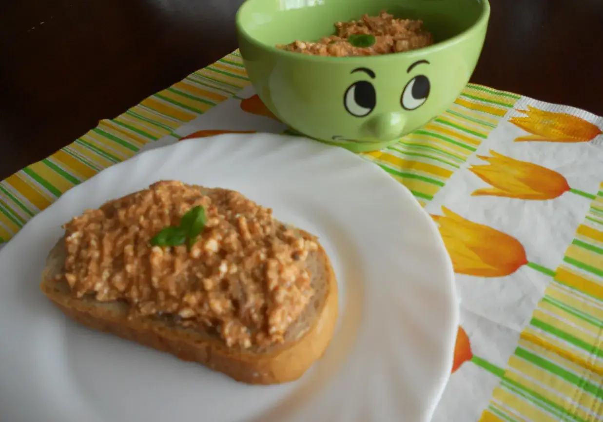 Pyszna pasta z makreli i twarogu na kromce chleba, udekorowana listkiem bazylii. Obok miseczka z pastą i uśmiechnięta buźka.