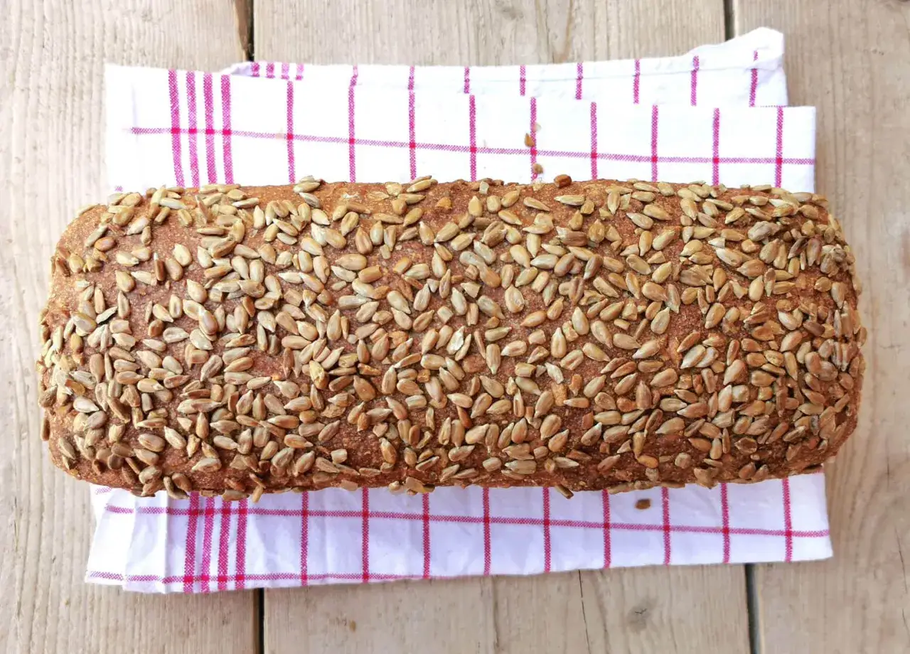 Ein rustikales Brot, bedeckt mit vielen Sonnenblumenkernen, liegt auf einem karierten K&uuml;chentuch.