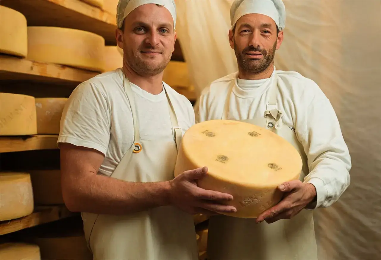 Due uomini con cappelli e grembiuli bianchi tengono una grande forma di formaggio. Dietro di loro, scaffali pieni di forme di formaggio. Forse un giorno scopriranno come fare il latte senza lattosio.