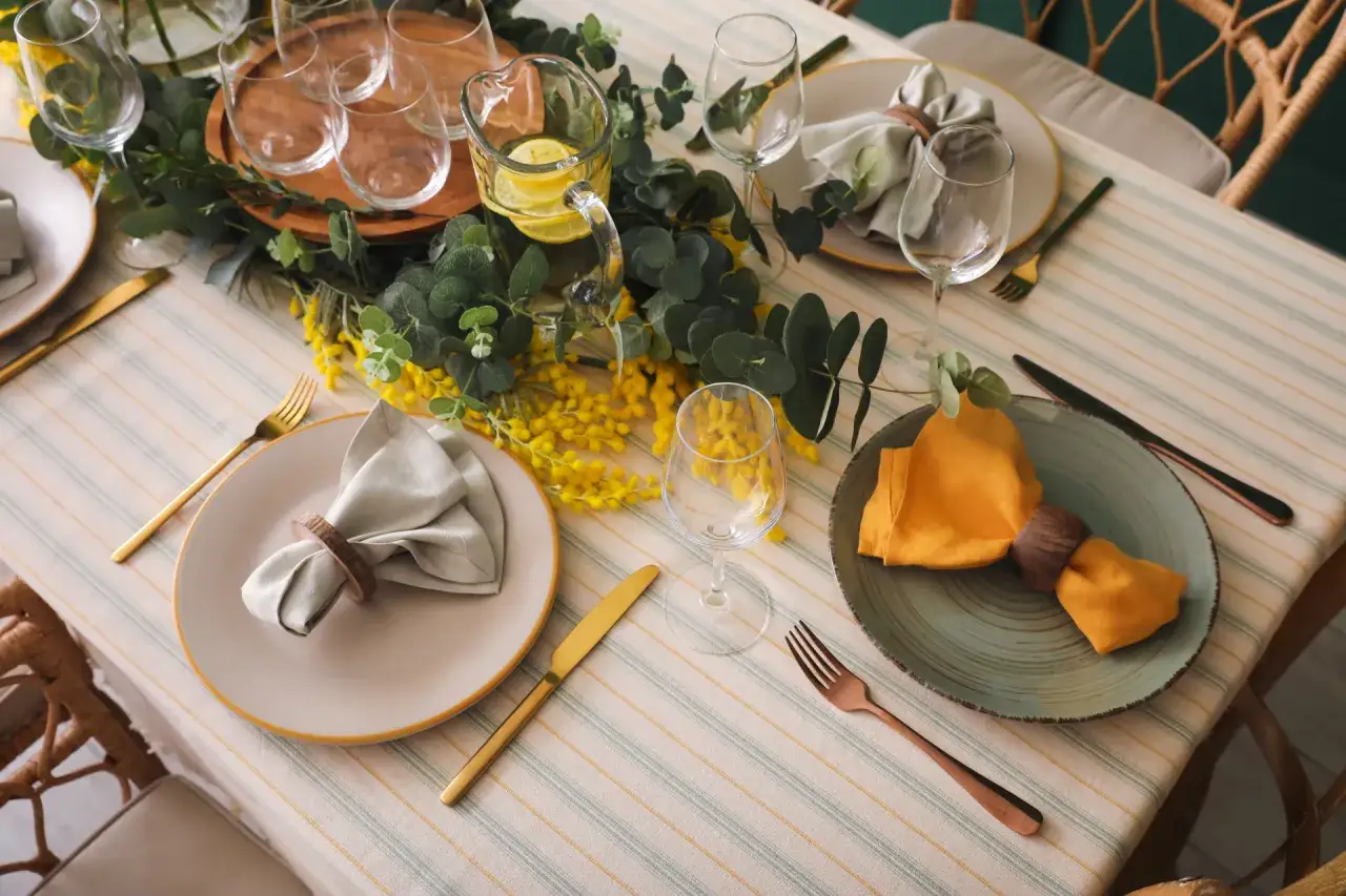 Une table joliment dressée pour un déjeuner d'anniversaire, avec des fleurs jaunes, de la verdure et des assiettes élégantes.