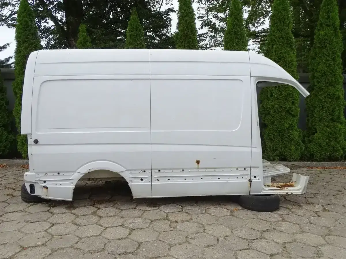 Biała, pusta karoseria Sprinter 906, bez kół i silnika, stoi na bruku, otoczona tujami.
