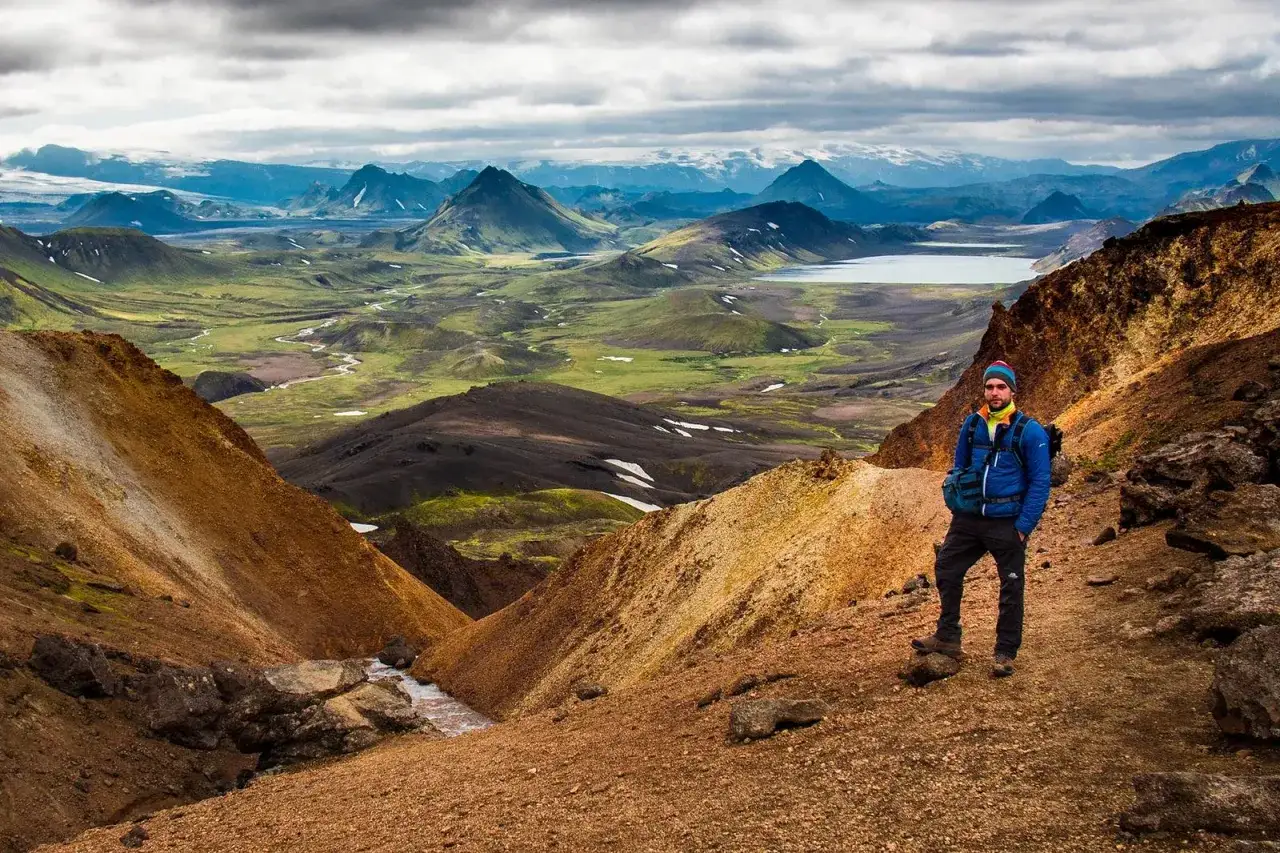A hiker stands on a ridge overlooking a vast, green valley with a winding river, part of the Laugavegur trail self guided.