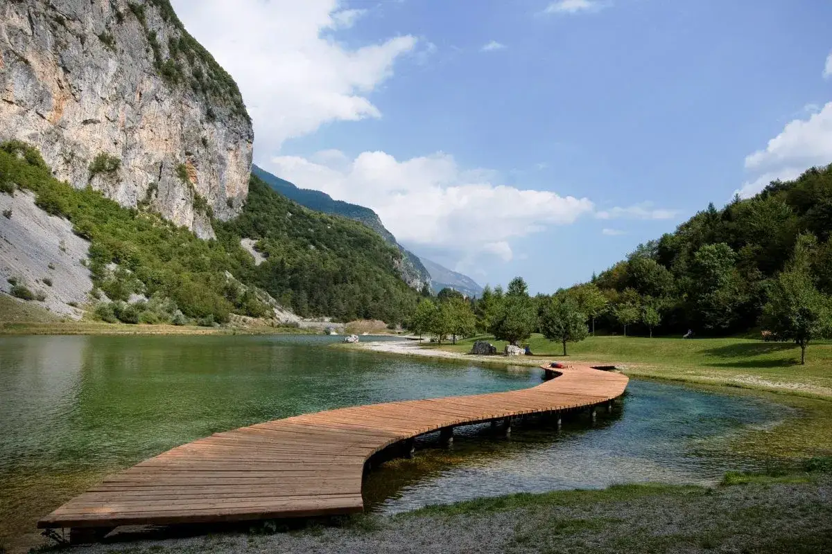 Un pontile in legno serpeggia sul **lago di Nembia**, circondato da montagne rocciose e verdi boschi sotto un cielo azzurro con nuvole bianche.