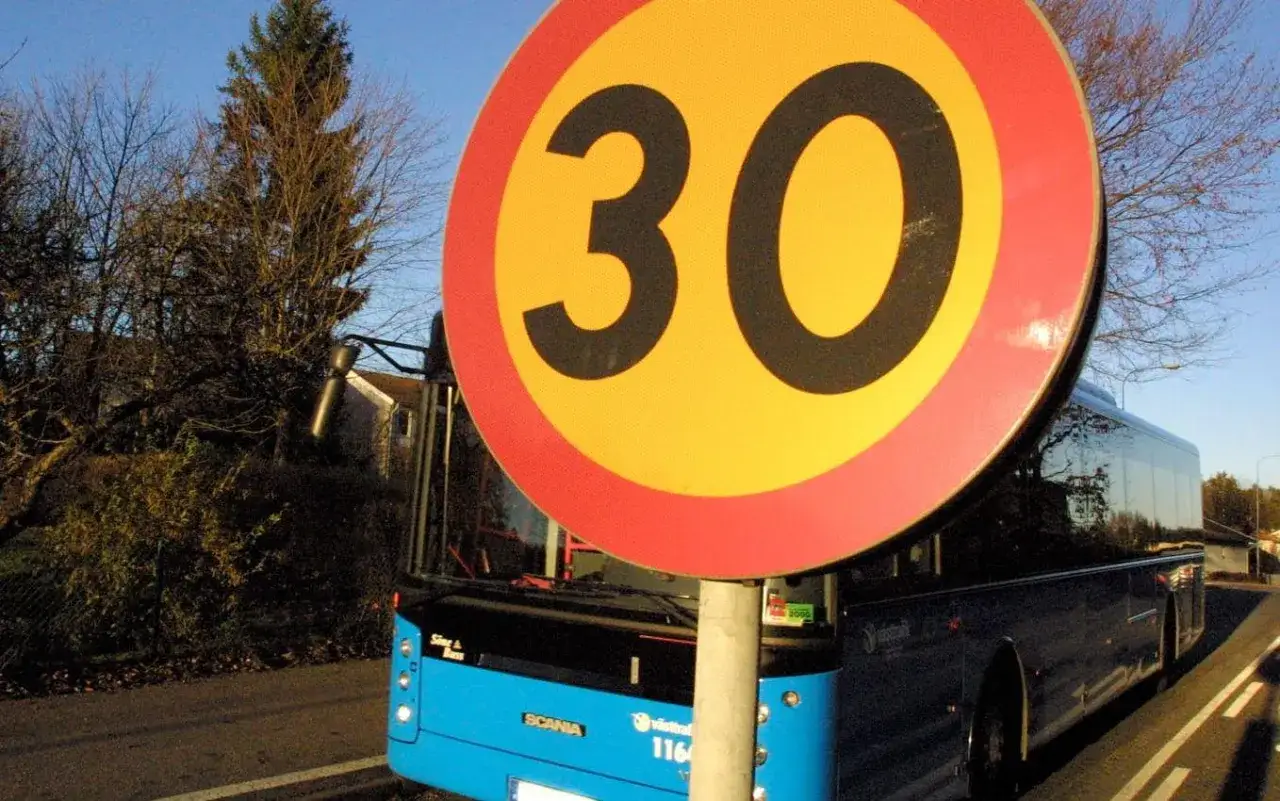 En gul skylt med röd kant visar siffran 30. En blå buss står parkerad bakom skylten.