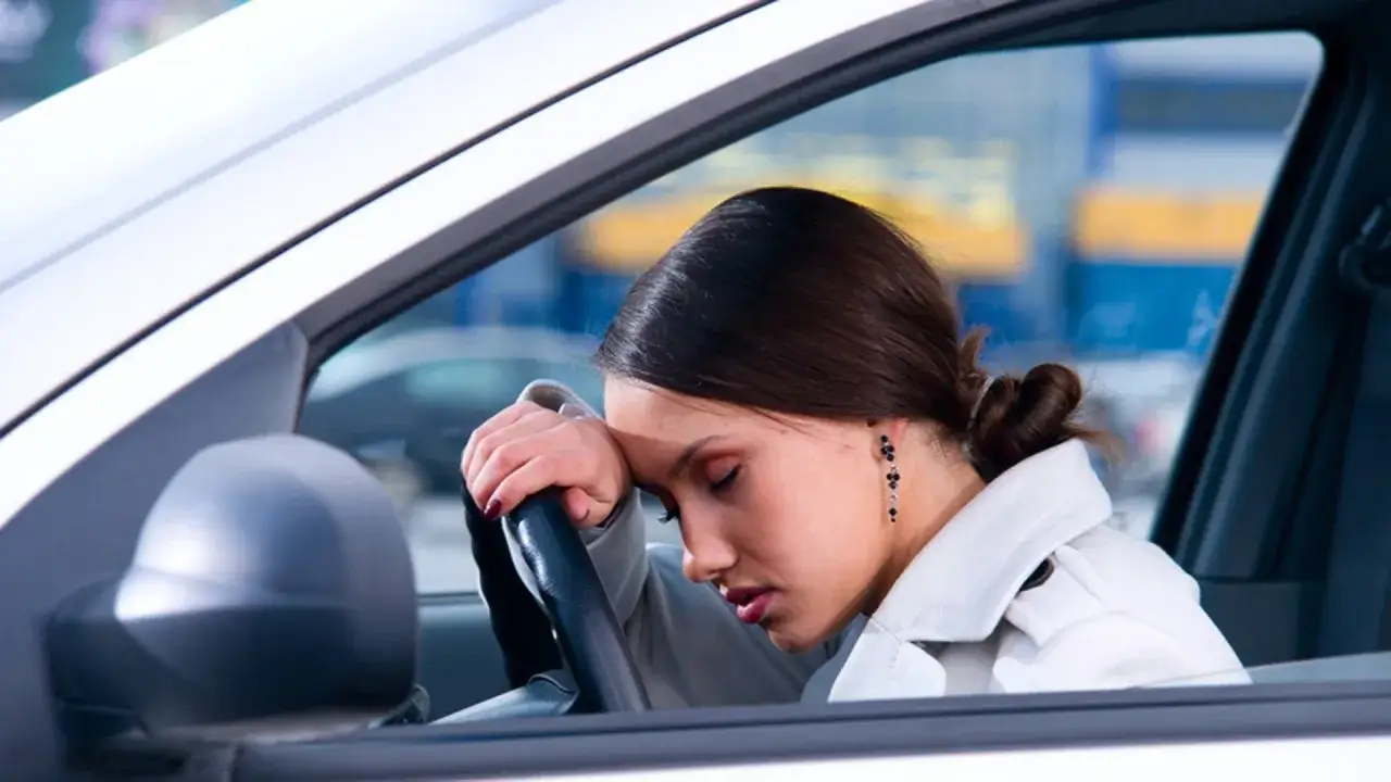 Donna esausta al volante, con la testa appoggiata sul volante. Un allarme variazione la spinge a fare una sosta.