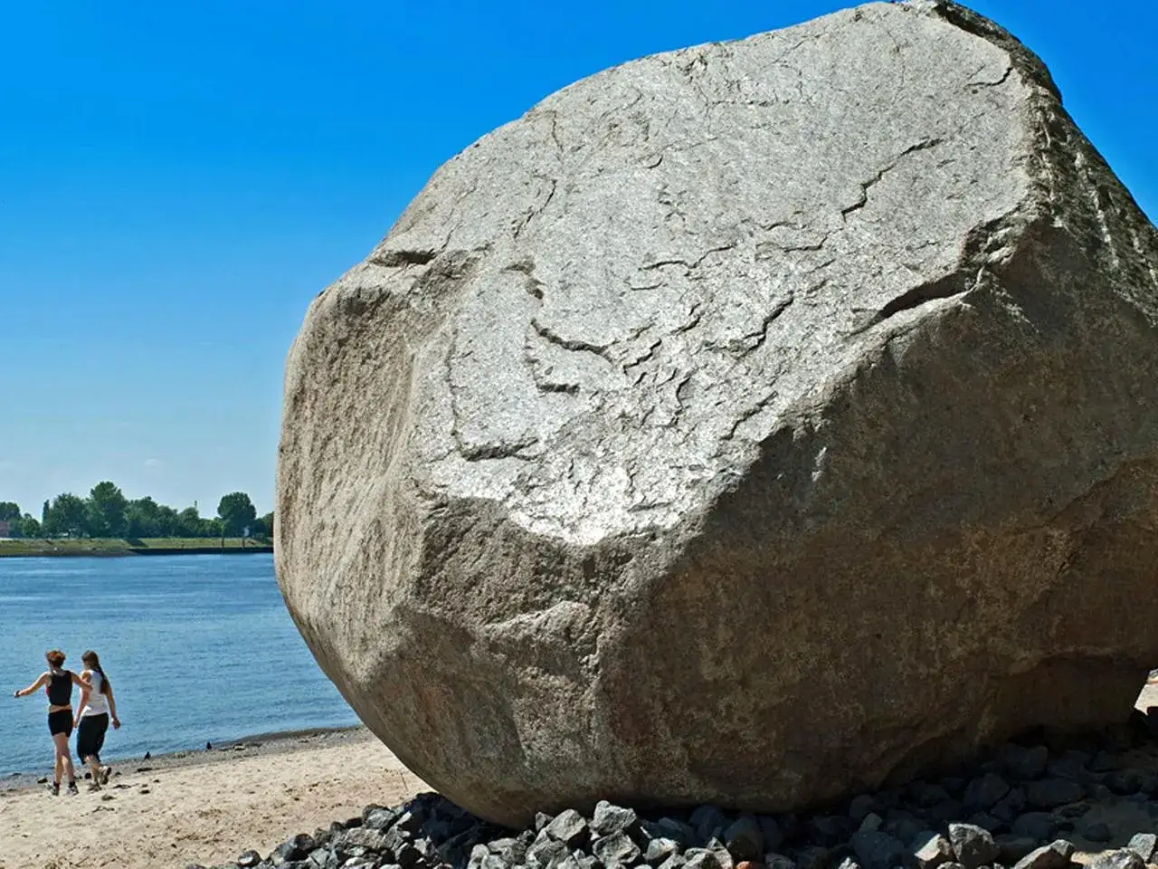 Zwei Frauen spazieren am Strand entlang, vorbei an einem riesigen Findling, dem 